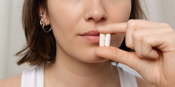 Nahaufnahme Gesicht einer jungen dunkelhaarigen Frau, die 2 Kapseln zwischen Daumen und Zeigefinger auf Höhe des Mundes hält