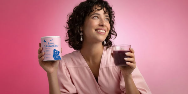 dunkelaarige Frau mit der Dose nu3 Luna Balance Elixir und einem Glas mit diesem Drink vor rosa Hintergrund